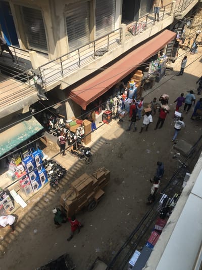 This image captures an overhead view of a bustling street market in Ojo, Nigeria. The scene is brightly lit by direct sunlight, creating distinct shadows across the unpaved, debris-strewn ground. Multi-story concrete buildings flank the market, featuring closed roller shutters on upper windows and a metal railing along an elevated walkway.
At street level, commercial activity is evident. A prominent, elongated reddish-brown awning shades a section of stalls displaying various electronics, including speakers, fans, and other boxed appliances. To the left, another section under a green awning showcases similar electronic goods. In the central-left area, numerous pairs of shoes and sandals are displayed on racks and piled directly on the ground.
Approximately 25 individuals are visible. Several men are actively transporting goods, carrying large cardboard boxes on their heads or in their arms. In the lower-center, a two-wheeled handcart is heavily loaded with stacked cardboard boxes, being maneuvered by two men. Other individuals are seen walking, standing, or observing the merchandise. A dense network of black electrical wires is strung overhead, connecting to poles and building facades.