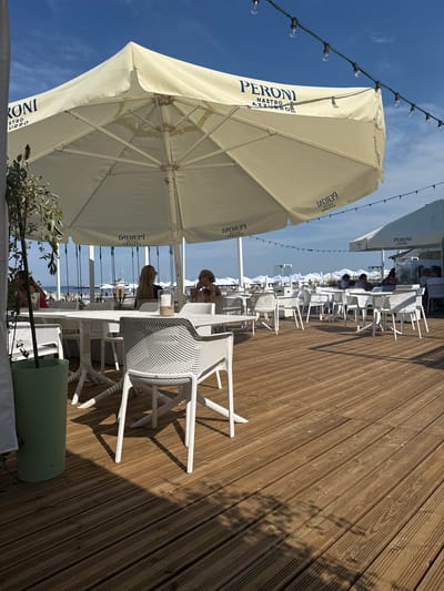 The image depicts a bright, sunny outdoor seating area at a seaside establishment in Sopot, Poland. A large beige umbrella with "PERONI NASTRO AZZURRO" branding provides shade over white tables and chairs arranged on a wooden deck. In the background, numerous other white umbrellas indicate a beach resort setting, with a glimpse of the Baltic Sea and a distant ship. Several individuals are seated at tables, engaged in conversation or enjoying the view. The scene is set on a clear day with a blue sky dotted with a few wispy clouds. The foreground features a large green planter with foliage, adding a touch of greenery to the scene.