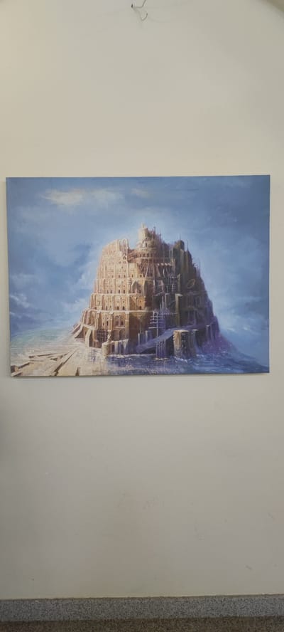 The image is a photograph of a painting hanging on a plain, textured white wall. The painting depicts a fantastical, towering structure reminiscent of the Tower of Babel. The structure is built of stone with many levels, arches, windows, and staircases, some of which appear unfinished or under construction. The scene is set against a dramatic sky with swirling blue and white clouds. The base of the structure appears to be surrounded by water or mist, with flat, stone pathways leading to it. The colors in the painting are muted, with warm earthy tones dominating the structure and cooler blues and grays in the sky and water. The photograph is taken from a straight-on perspective, with the bottom of the painting about a foot above a grey, speckled baseboard. The lighting is even, and there are no visible people or animals in the painting or the photograph. The image does not provide any information about the location in the city of Voskevaz, Armenia.