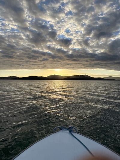 The image displays an open-water environment, likely at either sunrise or sunset, within the city of Dumbéa, France. The perspective is from the bow of a white boat, which occupies the lower foreground. A blue rope is secured to a metal cleat positioned on the foredeck of the boat, extending towards the lower right. The body of water exhibits a rippled surface, with a prominent, bright golden reflection of sunlight extending from the horizon towards the viewer. Along the horizon, a continuous line of dark-silhouetted land masses, appearing as hills or mountains, is visible. The sun, a strong golden light source, is positioned directly above the central part of these land masses. The sky is largely covered by a dense formation of textured grey and white clouds, with patches of clear blue sky visible through gaps in the cloud layer. No individuals or other vessels are present in the scene.