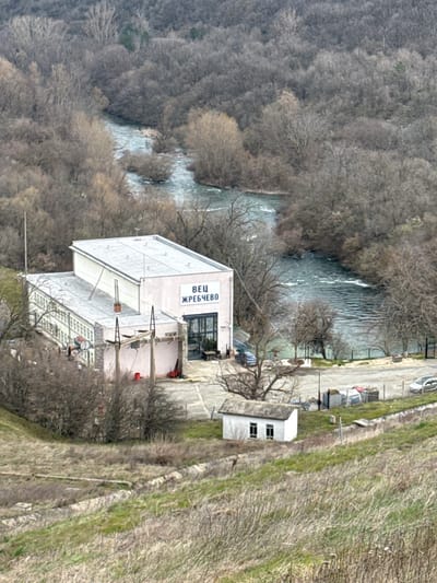An elevated, wide-angle image depicts the Zhrebchevo Hydroelectric Power Plant (ВЕЦ ЖРЕБЧЕВО) situated along a winding river, within the broader geographical area near Баня, Bulgaria. The central structure is a multi-story industrial building featuring a light pink facade with prominent blue Cyrillic text "ВЕЦ ЖРЕБЧЕВО," and a white facade with multiple windows, all under a light gray flat or low-pitch roof. External electrical infrastructure, including poles, lines, and equipment, is visible. Two vehicles, one dark blue and one lighter colored, are parked near the building's primary entrance. A smaller, white, rectangular outbuilding with a gray sloped roof and two frontal windows is located on a grassy slope below the main facility. The river, characterized by visible currents and small vegetated islands, flows through a valley densely covered by dormant deciduous trees. The immediate foreground consists of a steep, dry hillside with patchy green and brown grass and exposed soil. The scene is observed under an overcast sky. No individuals are present.