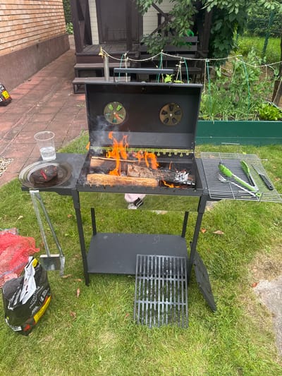 The image shows a backyard scene in the city of Orsongino, Russia. A black metal charcoal grill is in the center, with burning logs and flames inside. There is smoke rising from the grill. On the left side of the grill, on a black table, is a clear plastic cup with a clear liquid and a black plate with two pieces of grilled meat. To the right of the grill, a wire rack holds green-handled barbecue tools: tongs, a fork, and a knife. In the foreground, a black bag of charcoal and a red mesh bag are on the grass. A separate grill grate is placed on the grass near the grill. The background shows a brick wall, wooden porch, green plants, and a garden bed. The ground is covered with green grass and paving stones.