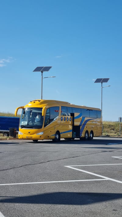 A bright yellow tour bus with blue accents is parked in an outdoor lot. The bus has its door open and a few passengers can be seen inside. There are two solar panels mounted on poles behind the bus. The bus's license plate reads "SMA 701". The scene appears to be in a rural or suburban area, with a grassy embankment and a blue tarp-covered structure visible in the background. The sky is clear and blue.