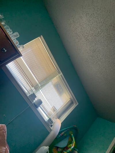 An upward-angled view captures an interior residential room, located in the city of Lancaster, United States of America. The left and lower wall areas are painted a solid teal, contrasting with a white textured popcorn ceiling occupying the upper and right portions of the frame. A large rectangular window, fitted with horizontal Venetian blinds that are partially closed, dominates the central and upper-mid sections, allowing bright daylight to filter into the room. Above the upper-left portion of the window, a dark brown wooden cabinet or shelf is partially visible, featuring a circular silver knob on its front. A row of clear plastic bottles with white caps is neatly aligned on top of this cabinet. A white flexible corrugated ventilation hose extends diagonally upwards from the lower-right corner, terminating near the window frame. Partially visible in the lower-right is a colorful infant activity mat or play gym. A white plastic jug is partially visible below the ventilation hose, and a light-colored, possibly upholstered object is visible in the lower-left corner.