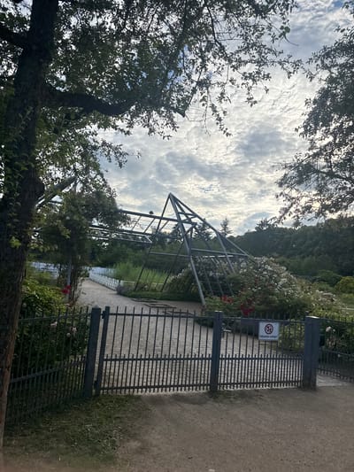 The image shows a view of a park-like setting with a metal gate in the foreground, partially obscured by trees. The gate is closed and has a sign with a red circle and a diagonal line over an image of a dog, indicating that dogs are not allowed. Text on the sign reads "Ballspielen verboten!" which translates to "Ball games forbidden!". Beyond the gate, there is a pathway and a trellised structure covered in roses, with white and red blooms. In the background, dense green trees form a backdrop against a sky filled with dramatic, cloudy formations. The lighting suggests it is daytime, possibly late afternoon, with the sun filtering through the clouds. The overall scene evokes a sense of a public park or garden, possibly in a German-speaking region given the text on the sign.