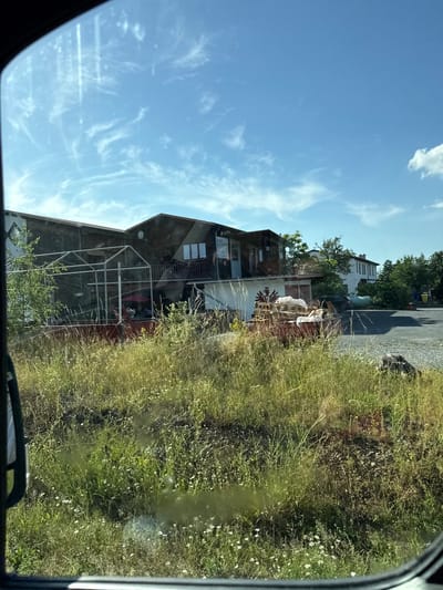 The image depicts a scene outside a window, likely from a vehicle, with the perspective rotated 90 degrees counterclockwise. The foreground is dominated by tall, dry grass and some wildflowers, suggesting an outdoor setting during a warm season. In the mid-ground, a multi-story building with a dark wooden exterior is visible. There's a metal structure resembling a cage or frame in front of the building, and several items are placed within it, including a pink object that could be an umbrella. On the side of the building, there's a dark red graphic with a stylized eagle and what appears to be text underneath.
Behind this building, another, lighter-colored building with multiple windows can be seen, along with trees and a parked car. The sky above is a clear blue with wispy white clouds, indicative of a sunny day. The overall impression is of a rural or semi-rural residential area in Germany, possibly during late afternoon given the lighting and the dry vegetation. There is no overt activity or event taking place, rather it's a snapshot of a static scene. A small amount of text is visible on the side of the building, but it's too blurry to be clearly read.