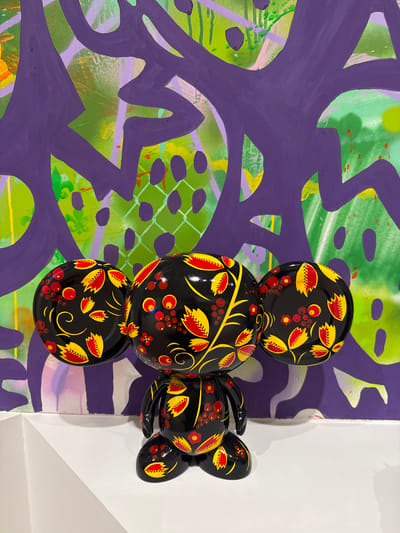 The image depicts a black, bear-like figurine with large, round ears, adorned with a traditional Russian Khokhloma painting pattern of red and yellow leaves and berries. The figurine stands on a white surface in front of a vibrantly colored abstract background. The background features large, sweeping purple shapes overlaying a green base with splashes of yellow, pink, and orange. The overall scene appears to be a display within an art gallery or exhibition space, possibly in Moscow, Russia, given the artistic style and cultural motifs.