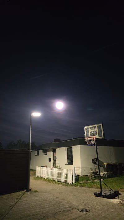 This media file presents a nocturnal outdoor scene in Ihringshausen, Germany, captured with a vertical orientation. The left half of the image is dominated by a dark night sky, across which faint, wispy light streaks are visible. A prominent, intensely bright, circular light source, likely the moon or a distant light, glows high in the upper-center, surrounded by a soft halo. Below it, a modern street light on a tall pole emits a powerful, elongated white light, illuminating the lower right portion of the frame.
On the right side of the image, a residential area is depicted. A white, multi-story building with dark windows serves as a backdrop. In the foreground, a white picket fence separates a grassy area or garden from a paved surface, which appears to be a street or pathway. This paved ground shows cracks and features a circular drain cover. A portable basketball hoop, characterized by a clear rectangular backboard and a red and blue net, stands on the paved surface, its dark base visible. Beyond the picket fence, a wire fence and some stacked items can be seen.
The scene is devoid of people or any discernible activity, conveying a quiet and still atmosphere. There is no visible text in the image. The primary light sources are the bright celestial body and the street lamp, defining the nocturnal setting.