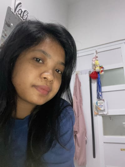 A selfie taken indoors shows a woman with dark hair and light brown skin. She is wearing a dark blue shirt. Behind her is a white wall with a window or paneled door. Above the window is a shelf holding various objects including what appear to be plush toys, blue ribbons, and a box or package. The location and city of Negara, Indonesia are not verifiable based solely on the details in the provided image.