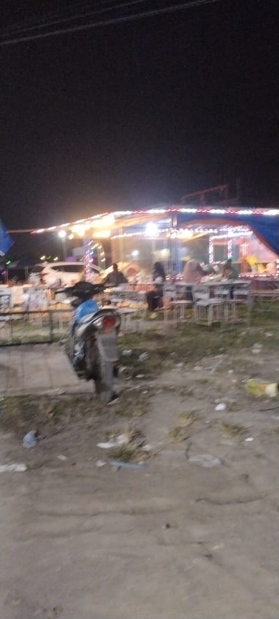 The image depicts an outdoor scene at night, likely a food stall or a small market in Gampongpaya, Indonesia. The setting is characterized by a tented structure illuminated by strings of festive lights, creating a warm and inviting ambiance. Several people are visible, some seated at tables, suggesting they are customers enjoying food or drinks. Others are standing or walking, possibly vendors or patrons moving through the area.
In the foreground, a scooter is parked, its blue and black body visible, adding a common mode of transportation to the scene. The ground appears to be unpaved and somewhat rough, with patches of dry grass and scattered debris, indicating an informal or temporary setup. The lighting from the stalls and the sky suggests it is nighttime, and the clear visibility implies fair weather.
The overall atmosphere appears relaxed and communal, with the lights contributing to a lively yet casual mood. The image captures a slice of local life, likely a popular spot for evening gatherings and dining. No visible text is present in the image.