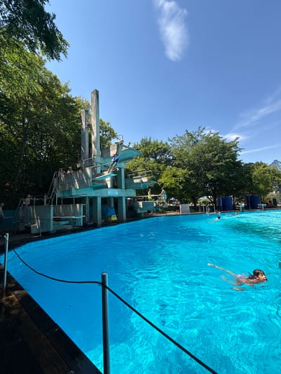 The image shows an outdoor swimming pool on a sunny day. The pool is filled with bright blue water and has wooden decking around a portion of it. In the background, a large diving platform made of concrete stands prominently, with several levels and ladders. Some people are visible on the diving platform, and one person is captured mid-dive. Others are in the pool, swimming. Lush green trees surround the pool area, and the sky is a clear blue with a few wisps of clouds. The scene appears to be located at the Freizeitbad in Rheinbach, Germany, an outdoor swimming facility with a diving tower.