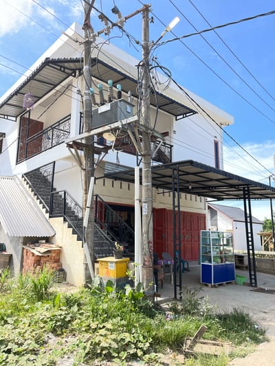 This is an outdoor, eye-level, medium shot of a residential building in an urban setting, likely in Dalam, Indonesia. The building is white with black accents, featuring multiple levels and an outdoor staircase with ornate black metal railings. A prominent utility pole with a complex network of electrical wires and transformers stands in the foreground, obscuring parts of the building. Various objects are visible around the property, including stacks of bricks, a metal display case with blue and glass panels, a green plastic container, and a purple birdcage hanging from an awning. Lush green vegetation grows alongside the building and concrete pathways. The scene appears to be from during the day, under a clear blue sky with some scattered clouds. No people are clearly visible. There is text on the utility pole: "0823 2281 7775".