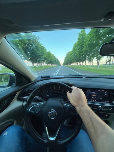 The image captures a first-person perspective from inside a car driving on a road lined with trees. The scene suggests a daytime journey, with bright sunlight and a clear blue sky visible through the windshield. The road appears to be a paved highway with white lane markings. A row of tall, leafy green trees flanks both sides of the road, creating a tunnel-like effect. In the foreground, a person's hands are visible on the steering wheel, indicating they are driving the vehicle. The interior of the car shows a dashboard with a digital display and a steering wheel with control buttons. The digital display shows the time as "21:00" and the temperature as "22". A song title "Красная краска" by "Ильгизар Хайруллин" is also displayed on the screen. The overall mood is one of a peaceful drive through a scenic, tree-lined route. The location is identified as Гарочыцкі сельскі Савет, Belarus.