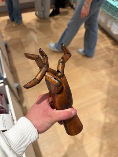A person holds a wooden articulated mannequin hand in an indoor setting. The hand is dark brown, with visible joints. The person holding the hand wears a white long-sleeved shirt. The background is a light brown floor, and out-of-focus legs wearing jeans and sneakers can be seen to the left. Other items on shelves are visible at the lower edges of the image. The image was taken at the IKEA location in Spreitenbach, Switzerland.