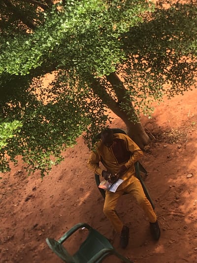 The image shows a man seated on a green plastic chair under a tree in Nteje, Nigeria. The man is wearing a brown and tan outfit and black shoes, and he appears to be reading a brochure or pamphlet. Another green chair is near him. The ground is red soil. The photo appears to have been taken from above.