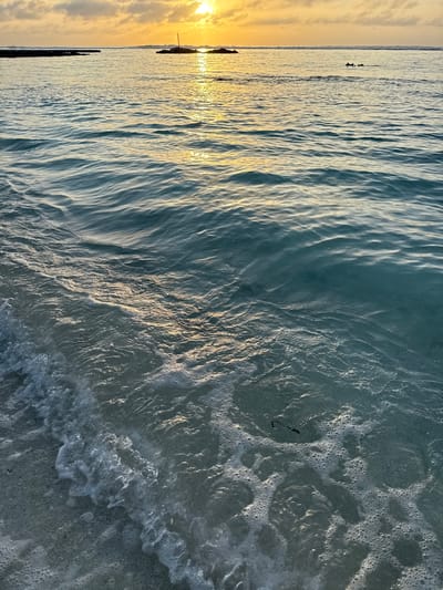 The image depicts a serene ocean scene at sunset, likely in the Maldives, given the characteristic turquoise waters and warm, hazy sky. The sun, a bright orb casting golden reflections on the water, is partially obscured by scattered clouds in the upper portion of the frame. The foreground is dominated by gentle waves lapping onto a sandy shore, creating a foamy, frothy edge. In the mid-ground, the water is calm with subtle ripples, and in the distance, a few dark shapes, possibly small boats or other marine life, are visible on the horizon. The overall impression is one of tranquility and natural beauty, indicative of a tropical paradise. There are no discernible landmarks or structures that would pinpoint a specific location within Malé.