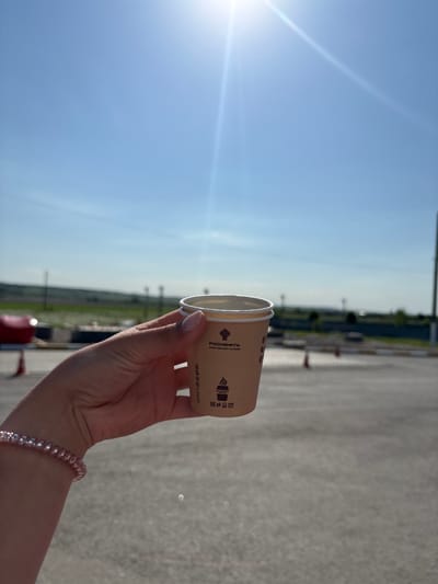 A person holds a brown paper coffee cup in their right hand against a bright blue sky. The cup features printed branding in Cyrillic script along with an image of a steaming coffee cup. A pink beaded bracelet adorns the person's wrist. The background shows an out-of-focus asphalt parking lot with distant utility poles and green vegetation. The bright sun, with its associated lens flare, dominates the left side of the frame. The location is an outdoor area possibly in Mar'evka, Russia, based on the branding language on the cup.