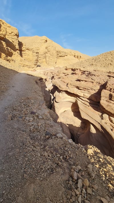 The image shows a narrow, winding slot canyon carved into light brown and beige rock formations. The canyon walls are smooth and sculpted, likely by water erosion. The bottom of the canyon is not fully visible, appearing dark and deep. Loose rocks and gravel cover the edges of the canyon, suggesting an unstable and arid environment. The sky is visible in the upper left corner, a clear bright blue. This is located in the city of Hevel Eilot Regional Council, Israel.