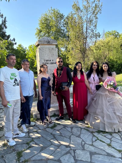 A group of seven people is posed for a photograph in Mihailovo, Bulgaria. The setting appears to be outdoors, likely a park or public area, with cobblestone pavement and a monument or stone structure in the background.
From top to bottom, the people are arranged in a line:
1. A woman in a light-colored, tiered dress holding a bouquet.
2. A woman in a light pink dress.
3. A woman in a red dress.
4. A man in a maroon suit.
5. A woman in a dark blue dress.
6. A man in a striped shirt and jeans.
7. A man in a white t-shirt.
The monument has Cyrillic text on it. There are trees visible in the background, and the sky is clear.