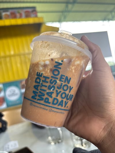 A person's hand holds a clear plastic cup containing a cold beverage, likely an iced coffee or tea, characterized by light brown liquid and swirling white, creamy components. Visible condensation on the outside of the cup indicates its chill. Blue text is printed vertically on the cup, reading "ENJOY YOUR DAY." and "MADE WITH PASSION." Below these phrases, smaller text appears to be social media handles: "@despresso_samalanga" and "@despressocoffee".
The scene is set outdoors or in a semi-outdoor commercial environment in Namplohbaro, Indonesia, under bright daylight. The background is blurred, showing yellow vertical slats, possibly from an awning or a building facade, along with indistinct signs and structures. The primary activity depicted is the act of holding the refreshing drink. The bright illumination suggests it is daytime, and the cold beverage implies warm weather. No specific emotions are directly conveyed by the limited visible subject matter.