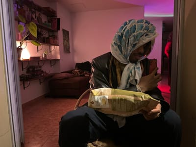 A person wearing a blue and white patterned headscarf, a dark jacket, and dark pants is seated in a wicker chair. They hold a bag of food in their left hand and a red smartphone in their right hand. The person appears to be taking a selfie or looking at their phone. The scene is a reflection in a mirror. The background includes a room with a brown sectional couch, shelves stocked with items, and a tile floor. In the mirror's reflection, a person wearing a red shirt is partially visible in a hallway. The room is lit by a lamp on the shelf and a purple light emanating from the hallway in the reflection. There is no evidence to indicate the location is in Montreuil, France.