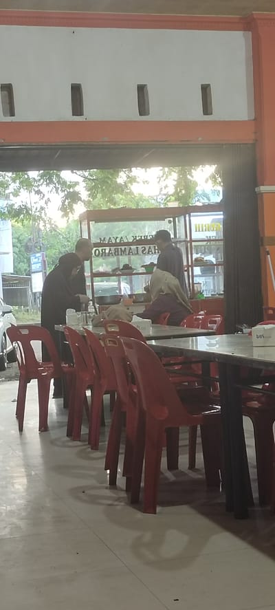 The image depicts an outdoor dining area, likely at a food stall or restaurant, in Lambaro, Indonesia. The setting appears to be illuminated by artificial lighting and natural daylight filtering through an opening above. Red plastic chairs are arranged around tables, some of which are covered with a white tablecloth and have napkins on them. In the background, a food display counter is visible, containing food items and a large pan. Three individuals are present: one person in a black hijab and dark clothing appears to be interacting with the food counter, another person in a light-colored hijab and maroon clothing is seated at a table, and a third person, a male in dark clothing, is standing and facing away from the camera near the food counter. The environment includes trees and foliage visible through the opening, suggesting an outdoor or semi-outdoor location. The overall scene suggests a casual dining establishment.