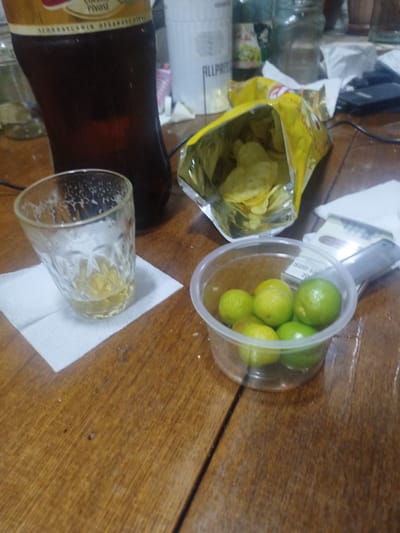 In the city of Sabunchu, Azerbaijan, a wooden table is set with various items. A large, dark brown bottle, possibly containing a beverage, stands prominently. Next to it, a clear glass with a textured pattern holds a small amount of amber liquid and some white foam residue. A white paper napkin is placed beneath the glass. To the right, an opened bag of potato chips spills its contents onto the table. In a clear plastic container, several small, green limes are gathered. In the background, out-of-focus objects include white packaging, a dark electronic device, and other glass containers. The lighting suggests an indoor setting with a warm, slightly dim ambiance.