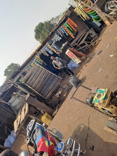 The image depicts a sunlit roadside commercial area in the city of Itobe, Nigeria. The scene is significantly tilted counter-clockwise. Two open-front stalls, constructed with corrugated metal walls and roofs, are situated on the left side of an unpaved path.
The stall on the left displays bundles of white material outside and hanging packaged goods within its entrance. An adult male, wearing a light-colored t-shirt and dark trousers, stands near the entrance of this stall.
The adjacent stall to the right features numerous colorful packaged goods hanging across its frontage. Inside, stacks of multicolored vehicle tires and several silver-colored wheel rims are visible. A brown A-frame sign stands in front of this stall. A second, partially obscured individual is visible deeper within this stall.
In the immediate foreground, the handlebars and front section of a red and blue motorcycle are visible, oriented away from the viewer. The unpaved ground in the mid-ground and foreground contains various objects: a green and yellow engine unit, a smaller engine or pump, a black garbage bag, and several metal reinforcing bars. A yellow jerrycan is also present on the ground near the left stall. The background consists of sparse trees under a clear sky, indicating direct sunlight.