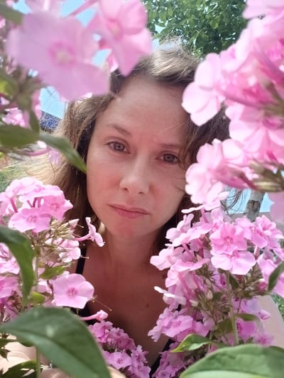 A woman is captured in a close-up, selfie-style shot, her face partially obscured by clusters of pink phlox flowers. She has light brown hair and is looking directly at the camera with a neutral expression. The background suggests an outdoor setting with visible greenery and a blue sky, consistent with a park or garden in Maslova Pristan', Russia. The lighting indicates it is daytime.