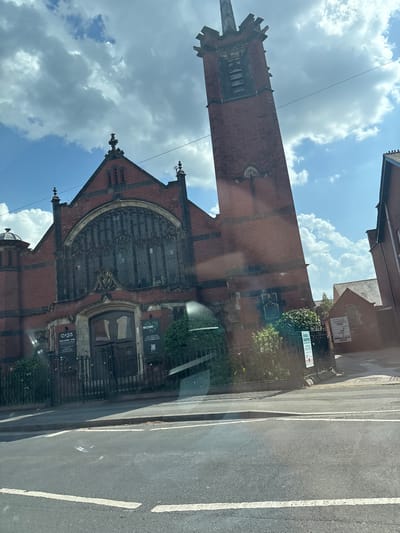 The image shows a street-level view of the exterior of the building, the Oasis Nottingham. A tall, red-brick tower with a detailed apex stands prominently against a blue sky filled with fluffy white clouds. The front facade of the building features a large arched window with intricate stone tracery. Black metal gates and fencing run along the perimeter of the property. In the foreground, a street with white lane markings is visible, suggesting the scene is taken from inside a vehicle. The building appears to be in Nottingham, United Kingdom, judging by the name of the building.