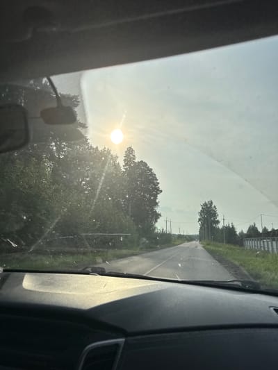 The image shows a view from inside a vehicle, looking out through the windshield towards a rural road. The sun is prominent in the sky, partially obscured by haze or dust, casting a bright glare. On either side of the road, there are trees and vegetation. Power lines and poles are visible along the roadside. In the distance, a white car is traveling on the road. The interior of the vehicle is visible in the foreground, including the dashboard. The scene appears to be in a temperate climate, likely during the daytime. The location is identified as Zarechnaya, Russia.