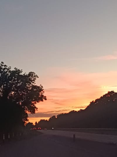 This is a photograph taken at dusk on a road in Arun, United Kingdom. The sky is a gradient of pale blue at the top, transitioning to soft pink and vibrant orange hues near the horizon. Clouds are visible, tinged with the colors of the sunset.

On the left side of the frame, a large, silhouetted tree with dense foliage dominates the foreground, its dark form contrasting with the bright sky. Along the edge of the road, a white post-and-rail fence is partially visible.

The road itself is dark asphalt, with lane markings faintly discernible. Several cars are driving on the road, their headlights and taillights illuminated, suggesting forward movement. One car's brake lights are particularly bright, indicating it is slowing down. In the distance, a motorcycle with its headlight on is also visible.

On the right side of the image, a dense line of trees forms a dark silhouette against the sunset, creating a wooded embankment or hillside. The overall atmosphere is serene, capturing the transition from day to night.