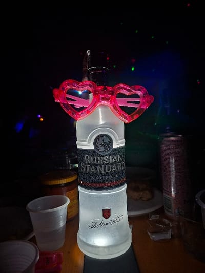 A forensic description of a dimly lit indoor scene in Kisyovtsi, Bulgaria, reveals a prominently displayed bottle of Russian Standard Vodka on a wooden surface. The frosted glass bottle is illuminated from below, causing it to glow, and features a black label with silver text and an eagle emblem. A pair of translucent pink heart-shaped novelty glasses rest on the bottle's neck. The dark background is dotted with numerous out-of-focus red, green, and blue lights, indicating decorative or festive illumination. To the left of the bottle, a translucent white plastic cup and a glass jar with a yellow lid are partially visible. To the right, a tall pink beverage can, a white paper plate holding a brown food item, and small pieces of debris are present on the table. No individuals are depicted.