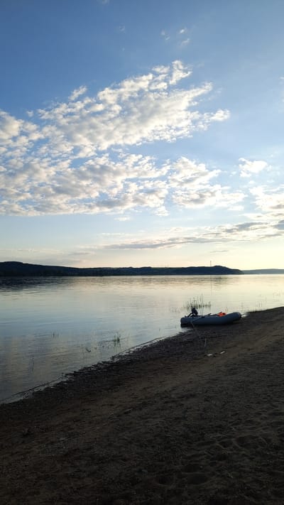 The image depicts an expansive body of water within or near Шолья, Russia, under a partially cloudy, blue sky. The upper portion of the sky is bright blue with numerous scattered white cumulus clouds, while the lower sky near the horizon is lighter, displaying a soft, warm illumination, indicative of either sunrise or sunset.
The water surface is calm, reflecting the sky and exhibiting minimal ripples, particularly near the shore. The far bank across the water presents a dark, continuous silhouette of densely wooded or forested hills, stretching horizontally across the entire frame. Towards the right side of this distant bank, a single transmission tower is visible, and further to the right, several tall, slender structures resembling wind turbines can be discerned against the horizon.
In the foreground, a dark, sandy and pebbled shoreline slopes gently down to meet the water. An inflatable boat, grey in color, rests partially in the shallow water and partially on the sand on the right side of the frame. An orange buoyancy aid or life vest is visible inside the boat. Adjacent to the boat, a fishing rod is propped upright, secured by a Y-shaped holder driven into the sand. Sparse, thin aquatic vegetation, resembling reeds or grasses, emerges from the shallow water along the immediate shoreline. There are no individuals present or engaged in any activity within the scene.