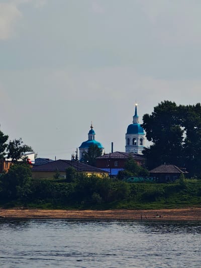 The image captures a view of the Yeniseysk Monastery in Yeniseysk, Russia, from across the Yenisei River. The monastery's distinct blue-domed churches and a tall bell tower are visible above a line of trees and houses situated on the riverbank. The water in the foreground reflects the overcast sky. Several small figures are present on the sandy shore, suggesting leisure activity. The overall scene depicts a tranquil, semi-rural setting with religious architecture dominating the skyline.