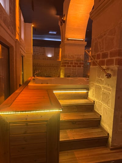 The image depicts an indoor scene in Ürgüp, Turkey, featuring a wooden staircase illuminated by LED strip lights. The staircase is surrounded by stone and brick walls and ceilings, giving the space a rustic or historical ambiance. A metal handrail with decorative ball fittings is also visible. Further elements include a white wall fixture, possible window openings, and glimpses into a dark space, suggesting the scene may be part of a building with both renovated and preserved historical features. There is no indication of any people within the frame. The orientation of the image is rotated, with the staircase appearing vertical in the frame.