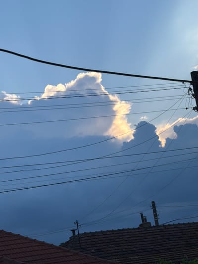 The image depicts a sky with clouds and a power line in Dalgopol, Bulgaria. The sky is a gradient of blue, with lighter shades towards the top and darker hues towards the bottom. Sunlight illuminates a large cloud formation, creating bright highlights and casting shadows on other clouds. Several power lines stretch horizontally across the frame, some appearing thicker and darker than others. A power pole with insulators and wires is visible on the right side of the image. In the foreground, the tops of tiled roofs of buildings are visible, along with chimneys. The overall scene suggests an outdoor, urban or semi-urban environment.