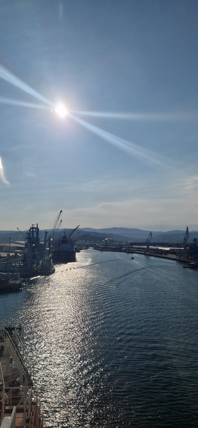 A wide, vertical, slightly low-angle, eye-level, full shot captures a bright, sunny day at the port of Ankaran, Slovenia. The sun is positioned in the upper left quadrant of the sky, casting brilliant rays across the scene and creating a strong glare on the water. The sky is a clear, light blue with a few wispy white clouds.

In the middle ground, a large cargo ship, identified by the name "MALCOLM" visible on its stern, is moving away from the viewer, leaving a wake of rippling water. To the left of the ship, a dock area is visible, featuring industrial cranes, smaller vessels, and what appear to be loading equipment. The water's surface is alive with reflections of the sun, creating a shimmering, textured effect.

The right side of the frame shows the expanse of the waterway, leading towards a distant shoreline. Along this shoreline, more industrial structures, including cranes and buildings, are visible, backed by rolling, tree-covered hills. The overall atmosphere is one of industrial activity under a clear, bright sky.