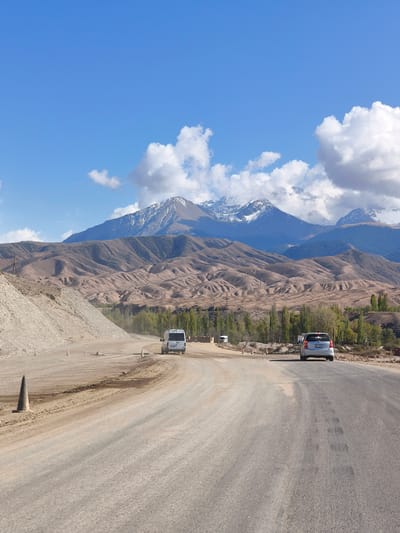 The image presents a vast, rugged landscape near Tamga, Kyrgyzstan, under a bright blue sky dotted with scattered white clouds. A wide, partially paved and partially dirt road dominates the foreground, curving gently to the right. The road surface appears dusty and uneven in sections. To the left of the road, a large pile of light-colored earth and rocks suggests ongoing construction or earthwork. Several orange traffic cones are visible along the roadside, indicating a temporary lane or work zone.

Two vehicles are traveling away from the viewer on the road. In the distance, a white panel van moves along a dirt section of the road. Closer to the foreground, a silver compact SUV or minivan is visible on the paved portion. Its rear license plate clearly displays "09KG 927 AEL," with "KG" indicating Kyrgyzstan.

The midground features rolling, barren hills, sculpted into soft, undulating forms, stretching across the scene in shades of brown and beige. A line of tall, slender green trees, likely poplars, grows between the road and these hills. The background is dominated by magnificent, snow-capped mountains, their peaks bright against the sky, indicating a high-altitude environment. The scene depicts a sunny day, suggesting fair weather for travel through this impressive natural setting.