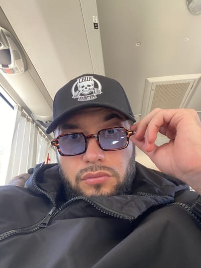 A man wearing a black baseball cap with a skull logo and brown-framed square sunglasses is taking a selfie. He is wearing a black hooded jacket with a zipper. He is indoors, possibly on a bus or train, indicated by the overhead compartment and window blinds. The scene is well-lit. The location is not specified in the image or its context.