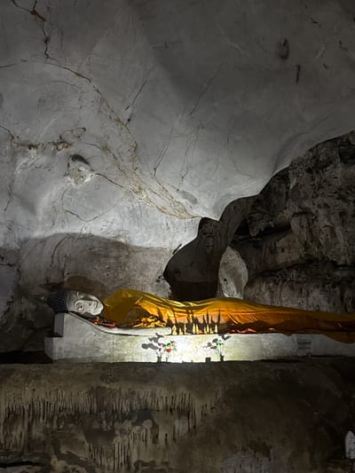 The image depicts a scene inside a cave. A large, light grey rock formation occupies the left side of the frame, displaying numerous cracks and fissures. To the right of this formation, a smaller, darker cave area leads further into the cavern. A concrete structure is seen in the center of the image, draped with a golden cloth. Attached to the structure is a head statue and some flower garlands. A large rock pillar occupies the right side of the frame. It appears to be inside the Tham Khao Luang cave, located in Ban Sahakon 2, Thailand.