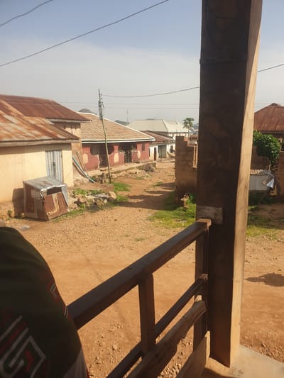 This is an image of a village scene, likely in Africa, not the city of Fresno, United States of America. The image shows a dirt road lined with several buildings with corrugated iron roofs. Some buildings are made of mud brick, while others appear to be made of concrete or stucco. There is a small, dilapidated metal structure on the left side of the road, possibly a shed or shelter. A person's arm and shoulder, wearing a green and red patterned garment, are visible in the foreground on the left, obscuring part of the scene. The sky is clear and blue with a few faint power lines visible. The overall impression is of a rural, somewhat impoverished setting.