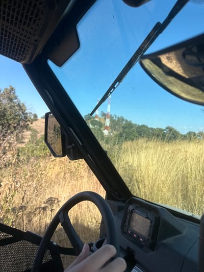 The view is from inside a utility terrain vehicle (UTV). A windshield wiper is visible against a clear blue sky. A red and white tower stands in the distance. The terrain outside is dry, grassy, and hilly. A side mirror is mounted on the vehicle's frame. The driver's hand rests on the steering wheel, with a digital display visible on the dashboard. The image suggests the vehicle is traversing an arid, outdoor environment.