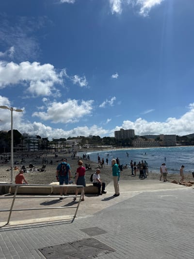 The image captures a beach scene in Peguera, Spain. A blue sky with scattered clouds dominates the left side of the frame. A beach lined with buildings and trees runs along the horizon, separating the sky from the blue water. People are on the beach, some swimming in the water. On the right side of the image, a gray brick walkway is visible, with several people standing and sitting along its edge, looking at the beach. A metal railing runs alongside the walkway.
