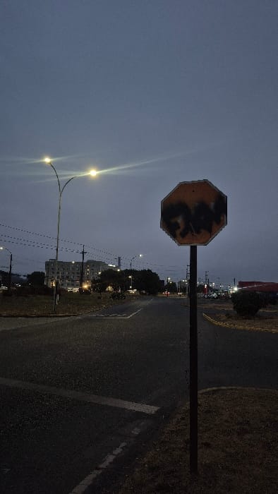A stop sign, obscured with dark markings, stands at an intersection near dusk. A dark road with a painted white line runs past, alongside an unpaved shoulder. In the distance, buildings are faintly illuminated by streetlights. Another lamppost arches above with two globes casting light. The sky is dark blue.