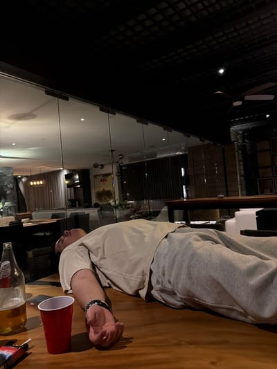 A person rests their head on a wooden table next to a red plastic cup, a partially full bottle of amber liquid, a cigarette, and a pack of cigarettes. The person is wearing a grey sweatshirt and a light-colored collared shirt, and a bracelet on their left wrist. The table is located near a glass wall, behind which there is a room with a television, artwork, and lighting fixtures visible. The location is in Santa Ana Yancuitlalpan (Chilhuacán), Mexico.