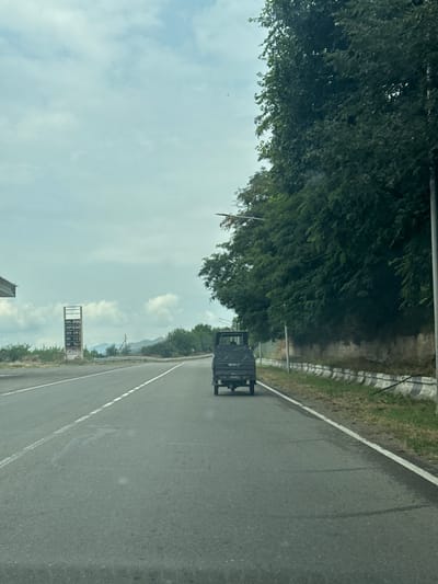 The image depicts a frontal view of a road in Rachisubani, Georgia, under a cloudy sky. A dark-colored, small utility truck is traveling away from the camera in the right lane. To the left of the road, a gas station canopy is visible, along with a price board displaying fuel costs. Further left, a line of trees and hills can be seen in the distance. The right side of the road is bordered by a concrete barrier and dense green trees. The road itself is asphalt, with white lane markings. The scene appears to be captured from a moving vehicle.