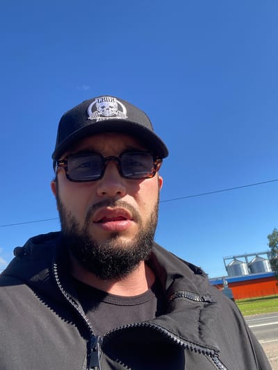 This media file is a close-up, angled selfie of a man against a bright blue sky. The man is wearing a black baseball cap with a white emblem on the front, sunglasses with patterned frames, and a black zippered jacket over a black t-shirt. He has a beard and his mouth is slightly open.

In the background, there are industrial silos, possibly grain elevators, painted with white and orange colors. A road and some greenery are also visible. The bright sky suggests it is daytime, likely sunny. The overall impression is of an outdoor setting, possibly in a rural or industrial area. There is no obvious activity or event taking place; it appears to be a casual portrait. No text is clearly visible in the image, though the emblem on the cap might contain text that is not legible. The location context provided is Kaykino, Russia.