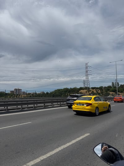 This media file shows a view from inside a vehicle, looking out at a street scene in Kâğıthane, Turkey. The primary focus is a bright yellow taxi, identified by the text "TAKSİ" on its side and the license plate "34 TFA 56". The taxi is one of several vehicles on a multi-lane road, suggesting a traffic situation. Another vehicle, possibly an orange car, is visible ahead of the yellow taxi.

In the background, a large electrical pylon and numerous power lines dominate the landscape, stretching across the frame and indicating a connection to infrastructure. Beyond the pylon, a green, tree-covered hillside is visible, with some buildings appearing in the distance. The sky is overcast with dramatic, swirling clouds, suggesting it might be a cloudy or possibly stormy day.

A portion of the filming vehicle's side mirror is visible in the bottom right corner, reflecting a person's hand holding a phone, suggesting the photo or video was taken using a mobile device. The overall scene depicts everyday urban or suburban life on a road with significant electrical infrastructure. The time of day appears to be daytime, given the illumination.