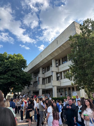 A large crowd of people stands in front of a tall, rectangular, light-colored building. A Bulgarian flag hangs on the building. Trees frame the building and crowd. The sky is blue with fluffy white clouds. The scene appears to be in Dulovo, Bulgaria. The crowd is densely packed, suggesting an event or gathering.